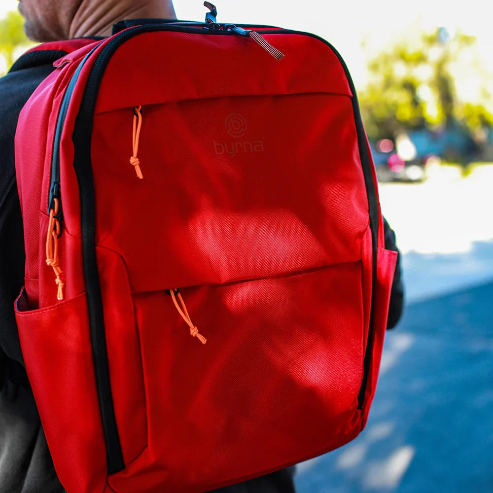 Red backpack with a Byrna logo on a blurred outdoor background.