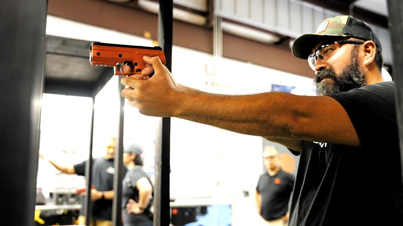 A bearded man wearing a black cap and glasses aims an orange Byrna SD pistol launcher in a shooting range. Other people are visible in the background through the range's glass partitions.