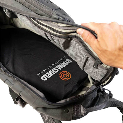 A close-up of a black tactical body armor shield inside a gray backpack, being opened by a hand. The shield features the words 'BYRNA SHIELD' and 'BACKPACK BODY ARMOR' printed in white, with the Byrna orange shield icon.